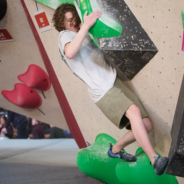 Iemand in een grijs shirt en een korte broek beklimt een indoor-bouldermuur, waarbij hij zich met zowel handen als voeten vastklampt aan groene en zwarte grepen, volledig geconcentreerd op zijn klim.
